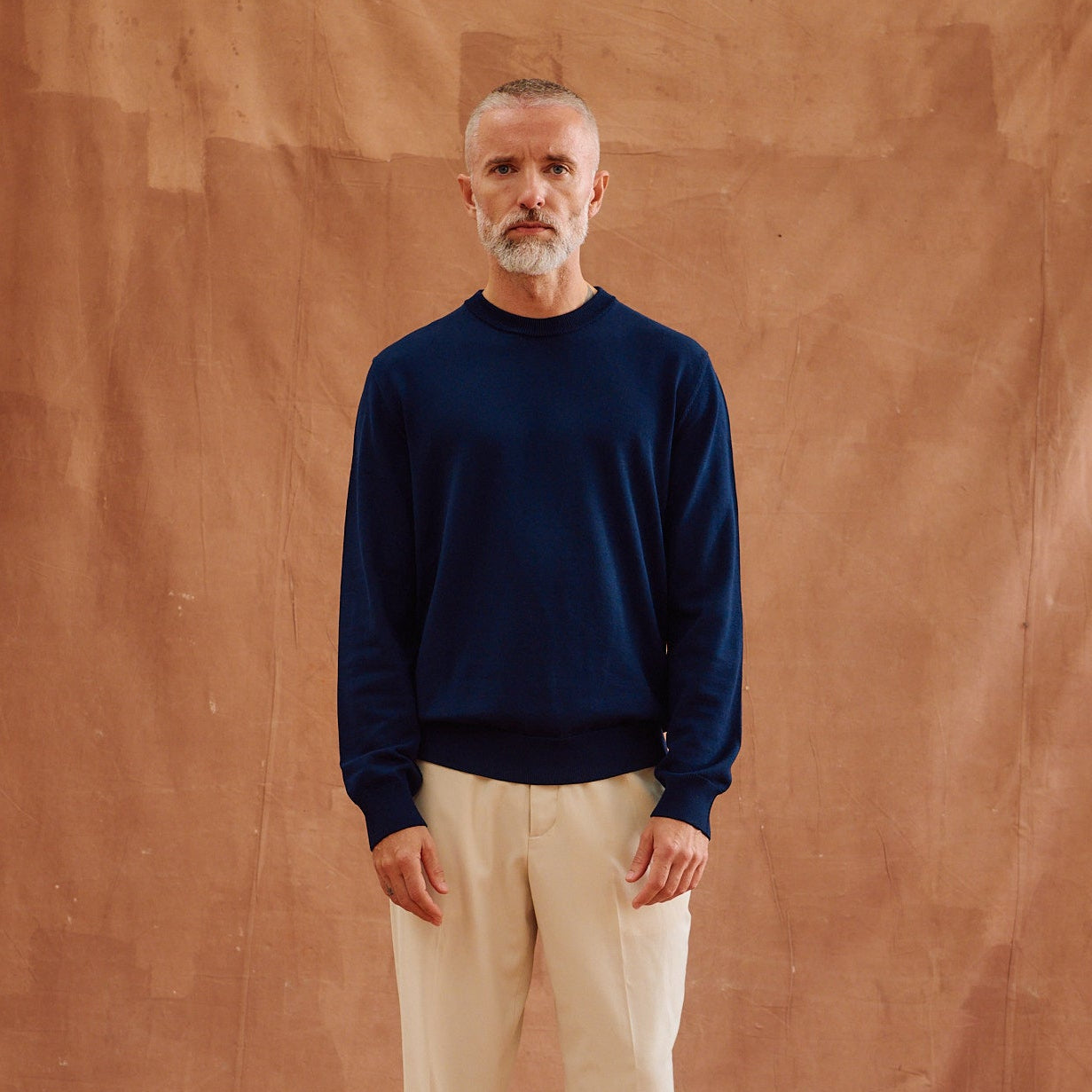 A man with a gray beard stands barefoot against a draped brown fabric backdrop, wearing a dark navy long-sleeve crewneck sweater and light beige trousers.