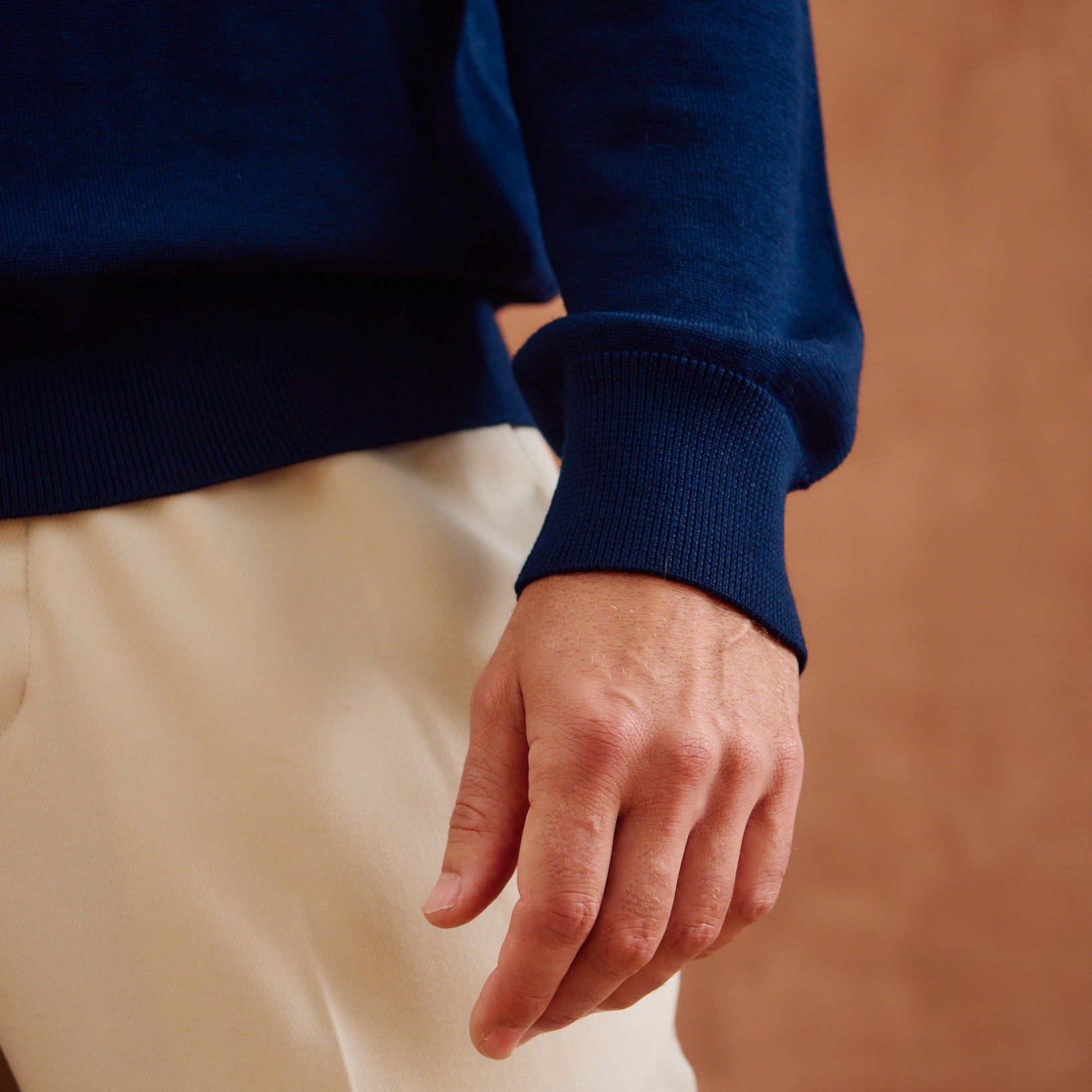 Close-up of a person wearing a navy blue ribbed knit sweater and cream-colored trousers, with the hand resting near the waist against a warm brown textured background.