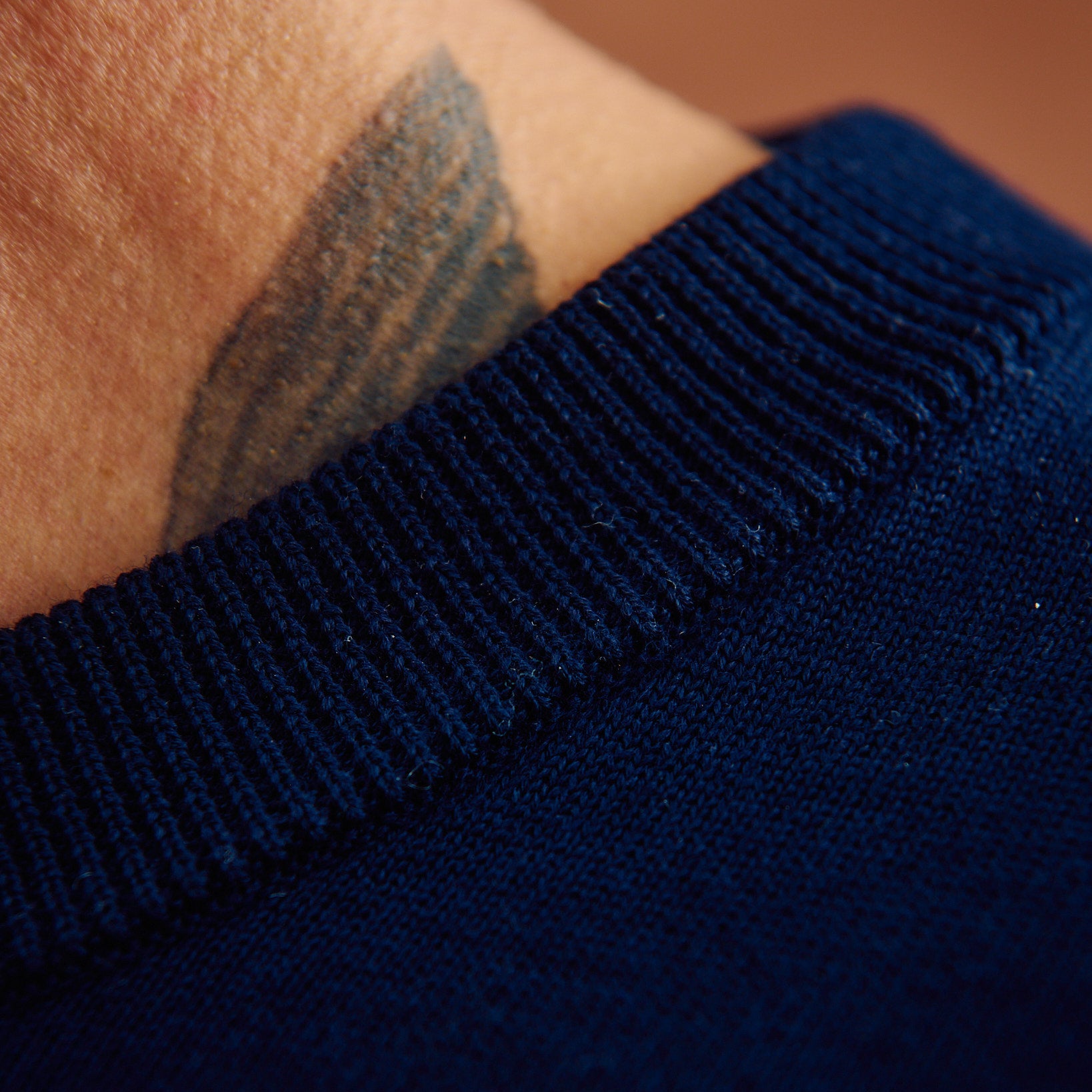 Close-up of a dark navy blue ribbed knit garment, likely a sweater, worn on a person's upper chest or shoulder, with skin and a tattoo visible at the collar.