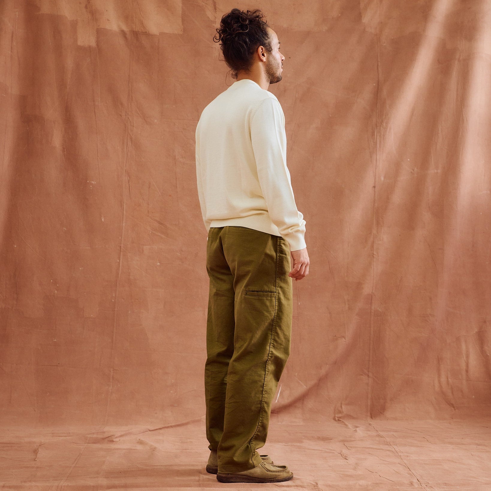 A man stands in profile wearing an off-white long-sleeve knit sweater and olive green cargo pants against a draped terracotta-colored fabric backdrop.
