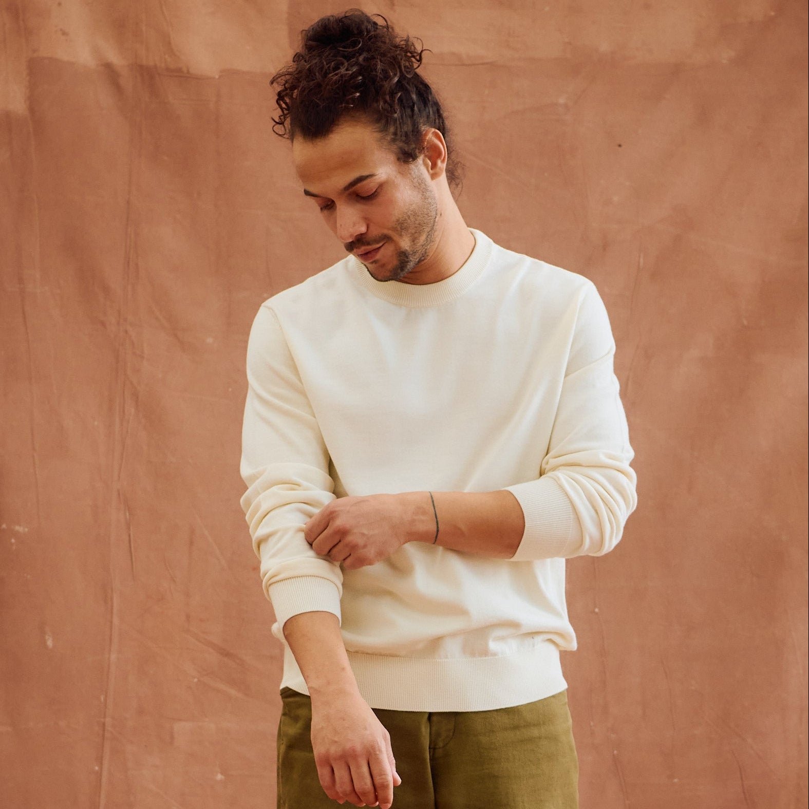 A man wearing a cream-colored crewneck sweatshirt and olive green pants, standing against a textured terracotta-colored wall.