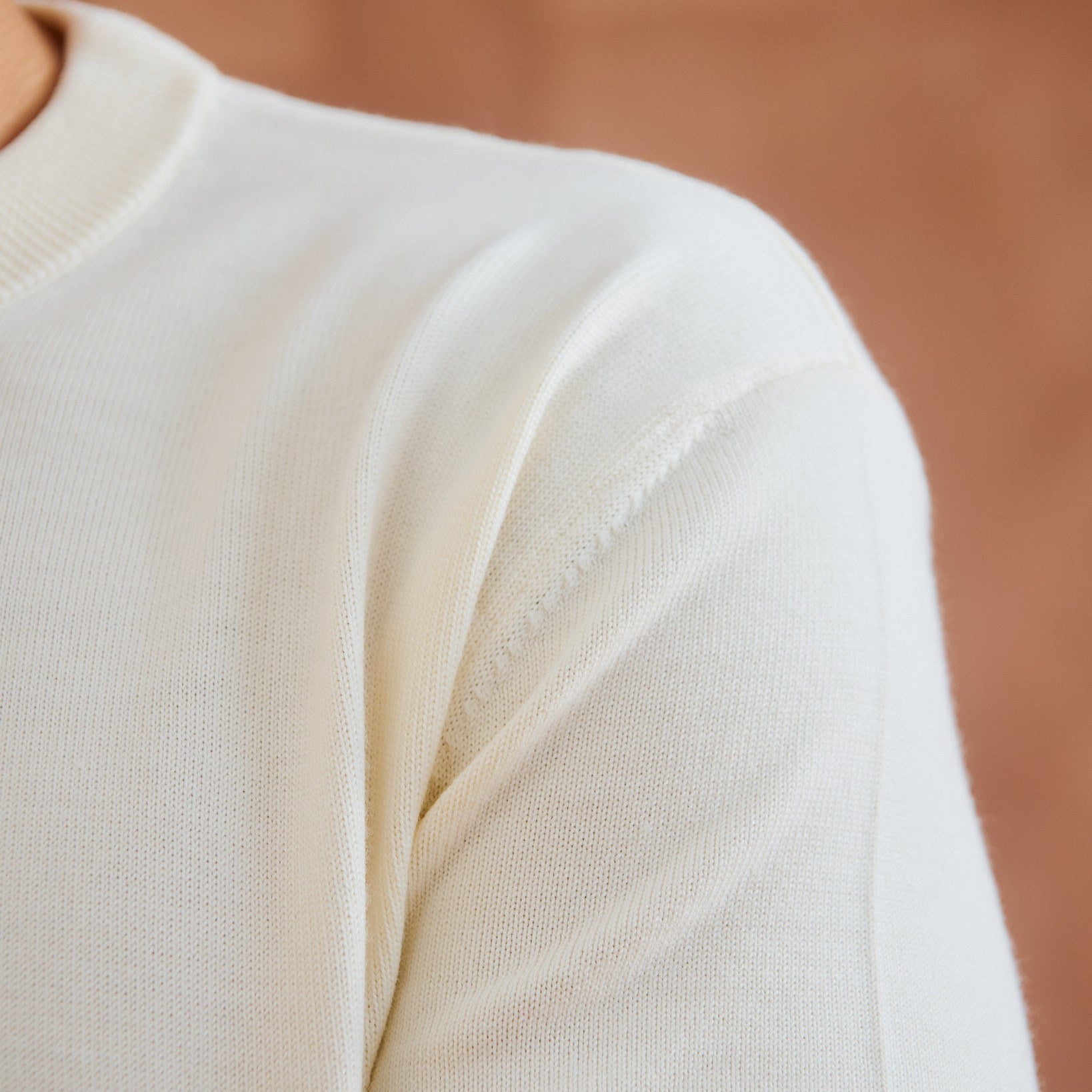 Close-up of a person wearing a cream-colored knit sweater, showing the shoulder and upper chest against a warm brown background.