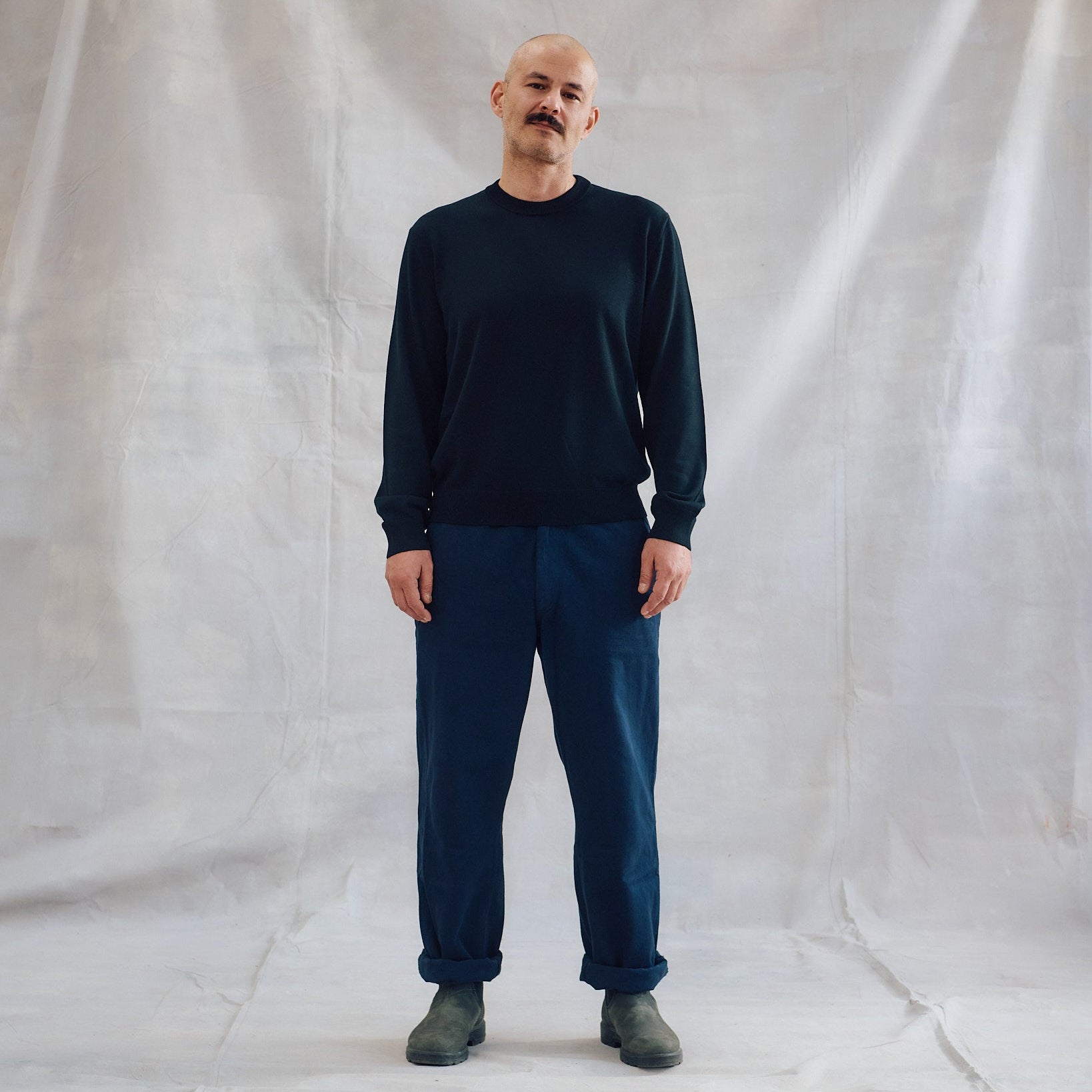 A man wearing a dark navy long-sleeve crewneck sweater and matching navy pants, standing against a draped white fabric backdrop.