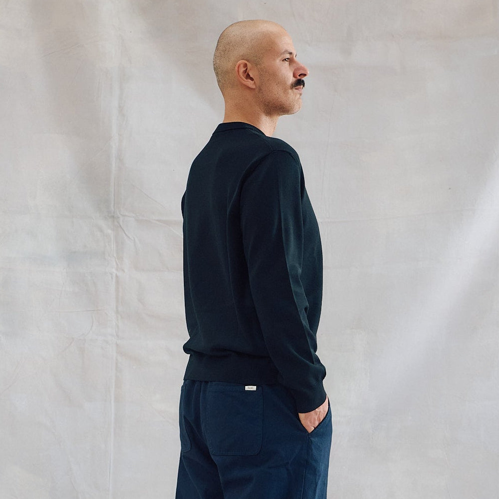 A man stands in profile wearing a dark black long-sleeve sweater, dark blue trousers, and dark gray boots against a draped white fabric backdrop.
