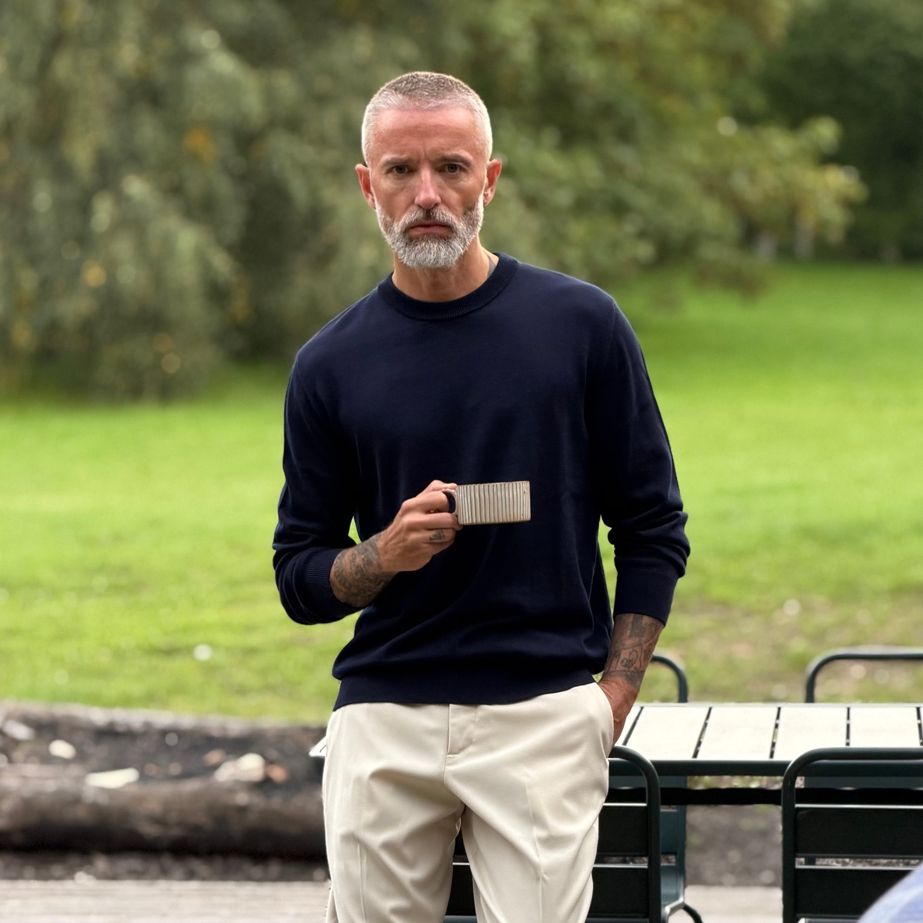 A man with a gray beard and tattoos stands barefoot on a wooden deck outdoors, wearing a dark navy long-sleeve knit and light beige trousers.