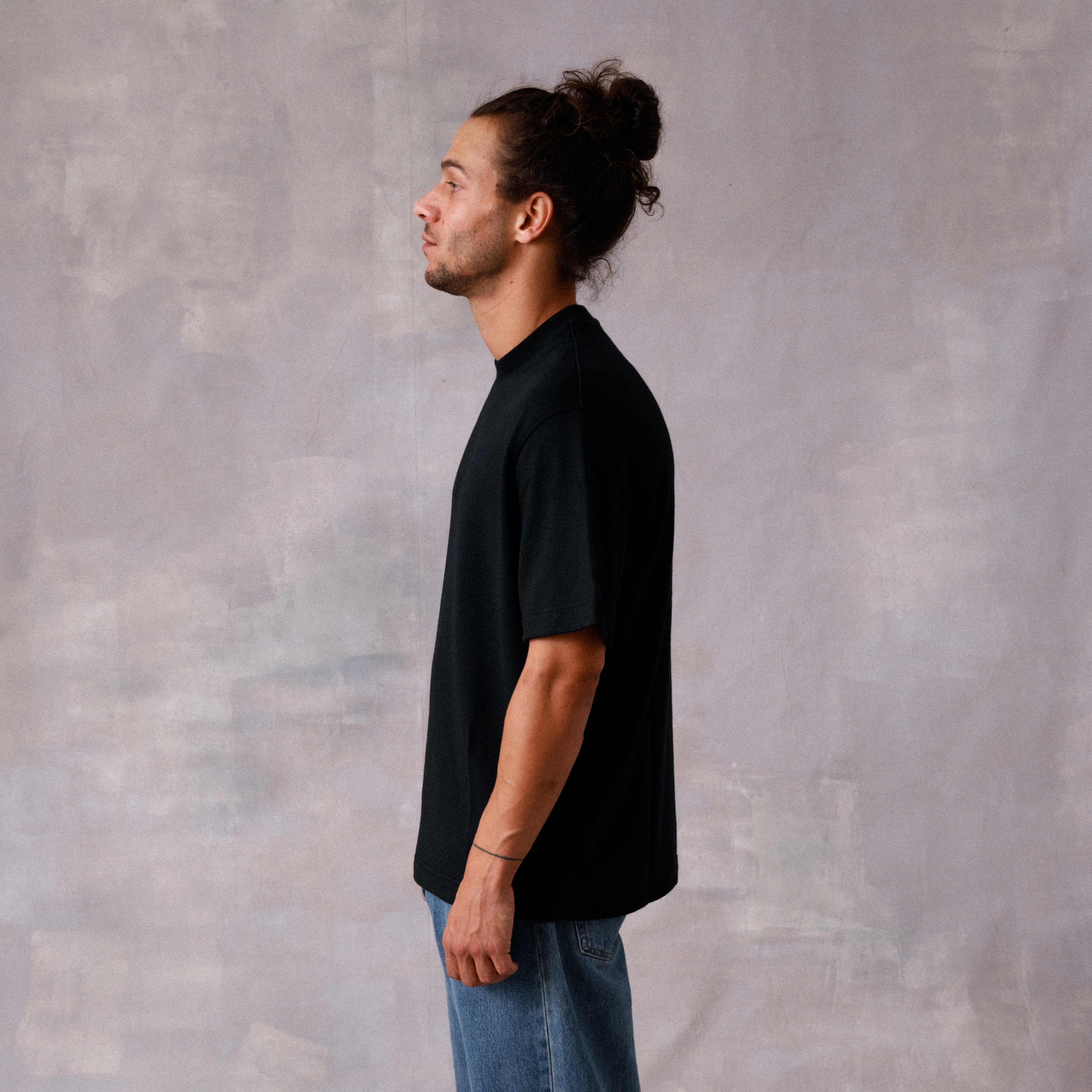 Man wearing a black t-shirt and blue jeans standing against a textured gray wall.